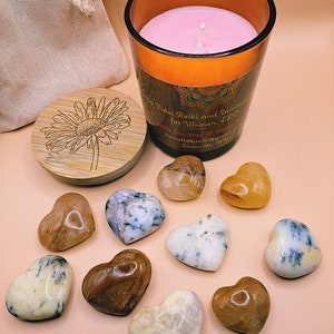 May include: A brown glass candle jar with a pink candle inside. The candle jar has a label that reads "Tides Reiki and Spiritual for Women, LLC 100% Soy Wax Hand Poured ringtidesreiki@gmail.com Louisville, KY".  There are nine heart-shaped stones in various colors and patterns, including brown, white, and clear.