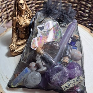 Puede incluir: Una bolsa de malla negra llena de varios cristales, piedras y pequeñas botellas. La bolsa está sobre una superficie de madera con una estatua dorada de una mujer en el fondo.