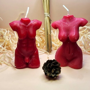 May include: Two red torso shaped candles, one male and one female, on a light pink surface with brown straw and a pine cone.