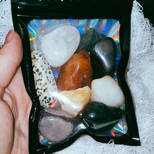 May include: A clear plastic bag filled with various tumbled stones, including white, brown, black, and grey stones.