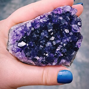 Può includere: Un grande ammasso di cristalli di ametista di colore viola scuro. Il cristallo ha la forma di un gruppo di punte e ha una texture ruvida e naturale.