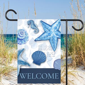 May include: A blue and white garden flag with a beach theme. The flag features watercolor illustrations of seashells, starfish, and a seahorse. The word "WELCOME" is displayed in navy blue. The flag is mounted on a black metal stand.