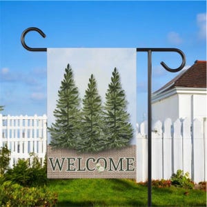 May include: A garden flag with three green fir trees on a light blue backdrop. The word "WELCOME" is in white on a burlap banner. The flag is held by a black metal stand in a garden with a white picket fence.
