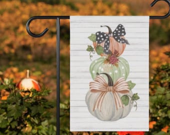 May include: A garden flag featuring a stack of three pumpkins in shades of orange, green, and gray, each adorned with a decorative bow. The flag is mounted on a black metal stand, perfect for fall decor.