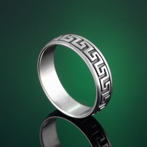 Könnte beinhalten: Ein silberner Ring mit einem griechischen Schlüsselmuster. Der Ring befindet sich auf einem grünen Hintergrund.