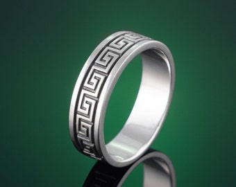 Peut inclure: Une bague en argent avec un motif de clé grecque. La bague est sur un fond vert.