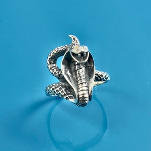 Puede incluir: Un anillo de plata con un diseño de cobra serpiente detallado. La serpiente está enroscada con la cabeza levantada y la boca abierta.