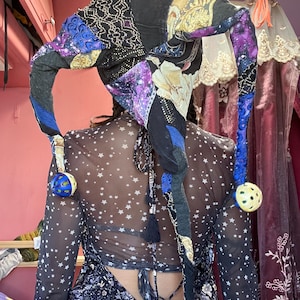 Könnte beinhalten: Ein farbenfroher Narrenhut mit langen, herabhängenden Spitzen. Der Hut hat ein Patchwork-Design mit schwarzen, lila, blauen und goldenen Stoffen. Jede Spitze endet mit einer kleinen, runden Glocke. Ein skurriles Accessoire.