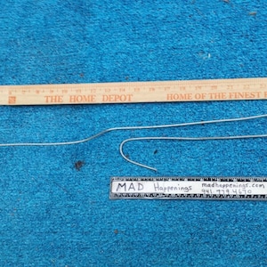 May include: A bent metal wire with a looped end and a smaller curved section. A wooden ruler with black markings and the text "THE HOME DEPOT" is above the wire. A small ruler with text "MAD Happenings" is below the wire.