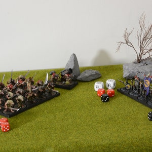 Op de afbeelding: Een tabletop-gamescene met twee legers die tegenover elkaar staan. Het ene leger bestaat uit kleine, bruine, rattenachtige wezens met zwaarden. Het andere leger bestaat uit grotere, humanoïde figuren met schilden en zwaarden. Er liggen verschillende rode en zwarte dobbelstenen op de tafel.