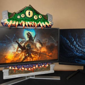 Op de afbeelding: Een op maat gemaakte monitorstandaard met een groen monsterontwerp, met meerdere ogen en tanden. De standaard staat bovenop een computermonitor, met een bijpassend ontwerp eronder. De monitor toont een fantasie-videogame scène.