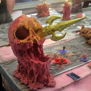 Op de afbeelding: Een 3D-geprinte schedel met een gouden speer die door de oogkas steekt. De schedel is dieprood en heeft een getextureerd, gesmolten uiterlijk. De speer heeft een drietandpunt en is versierd met een schedelontwerp.