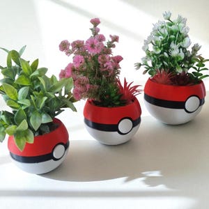 Può includere: Tre vasi rotondi rossi, bianchi e neri, simili a Poké Ball, ognuno con piante diverse. Uno ha foglie verdi, un altro fiori rosa e il terzo fiori bianchi. I vasi sono su una superficie bianca.