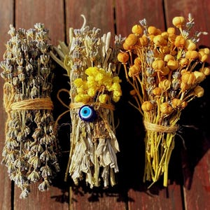 May include: Three bundles of dried herbs tied with twine. The bundles are lavender, white sage, and yellow dried flowers. A blue evil eye charm is attached to the white sage bundle.