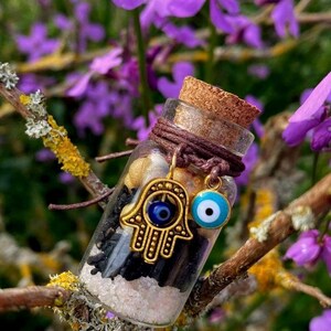 May include: A small glass bottle with a cork top filled with black sand, white crystals, and gold charms. The bottle is decorated with a brown string and two blue evil eye charms. The bottle is sitting on a branch with purple flowers in the background.