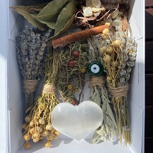May include: A white box filled with dried herbs, flowers, and a white heart-shaped selenite crystal. The box also contains a cinnamon stick, bay leaves, and a blue evil eye charm.
