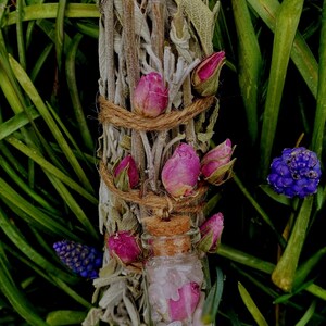 May include: A bundle of white sage smudge sticks tied with twine and decorated with dried pink roses. A small glass bottle filled with clear quartz crystals and a pink rose is attached to the bottom of the bundle.
