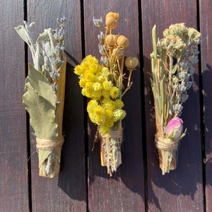 May include: Three bundles of dried herbs and flowers tied together with twine. The bundles are made with sage, lavender, yarrow, and other dried flowers. The bundles are arranged on a wooden surface.