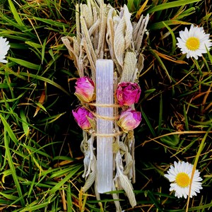 May include: A sage smudge stick with a clear crystal and dried pink roses, tied with twine. The bundle rests on green grass and is surrounded by white daisies with yellow centres.