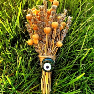 May include: Dried lavender bouquet tied with twine and topped with a blue evil eye charm. The bouquet is sitting on a bed of green grass.
