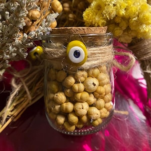 May include: A small glass jar filled with dried white flower buds, tied with twine and a blue and white evil eye charm. The jar is sitting on a pink surface.