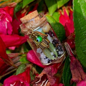 May include: A small glass bottle filled with various herbs, crystals, and a gold charm with a green eye. The bottle is sitting on a bed of pink flowers.