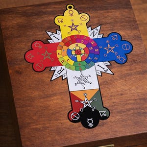 Pode incluir: Uma caixa de madeira com um design de cruz decorativa. A cruz apresenta um emblema circular colorido no centro, rodeado por símbolos e formas geométricas em vermelho, amarelo, azul e verde. A caixa tem um fecho de latão.