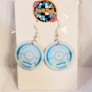 Puede incluir: Un par de pendientes colgantes circulares con un diseño azul claro, detalles holográficos y líneas concéntricas finas. Los pendientes están sujetos a una tarjeta blanca que muestra el logotipo Funky Finds en la parte superior.