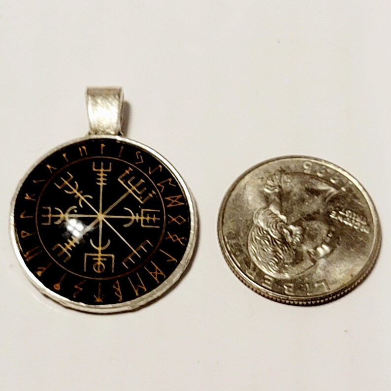 May include: A silver-toned pendant with a black face featuring a gold-colored Vegvisir symbol. The pendant is round and has a loop at the top for a chain. A quarter coin is next to the pendant for size comparison.