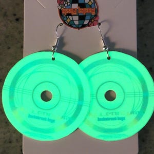 May include: A pair of circular earrings shaped like compact discs with a bright neon green glow. Each earring features the text "Backstreet Boys" printed in small letters near the bottom. The earrings hang from silver-toned metal hooks on a white card.