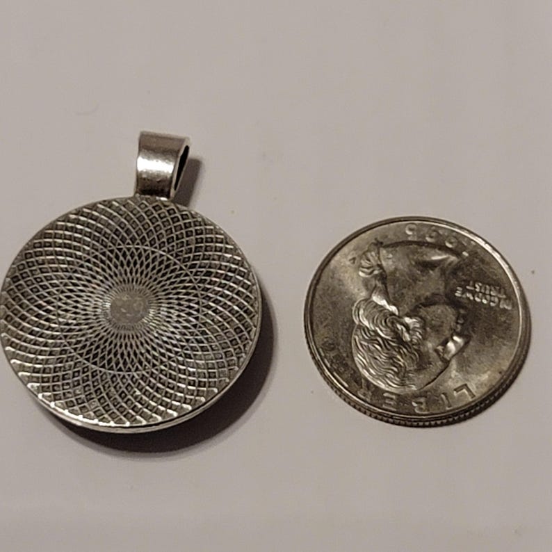 May include: A silver-toned pendant with a circular design and a loop for a chain. Next to it is a U.S. quarter coin for size comparison. The pendant has a detailed, textured pattern on its surface.