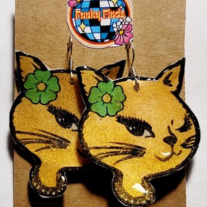 Può includere: Un paio di orecchini a forma di gatto con glitter dorati e fiori verdi. Gli orecchini sono su una carta marrone con un logo a forma di palla da discoteca che dice "Funky Finds".