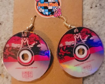 May include: A pair of earrings made from repurposed compact discs. The discs feature a red, black, and white design with a woman sitting on a creature. The earrings have gold hooks.