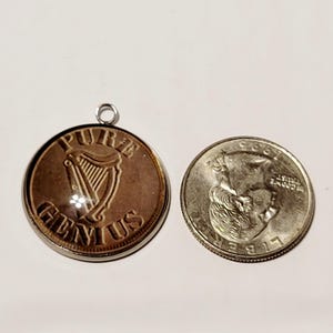 May include: A copper-colored pendant with the words "PURE GENIUS" and a harp design. Next to it is a silver quarter coin. The pendant has a silver loop at the top for hanging.