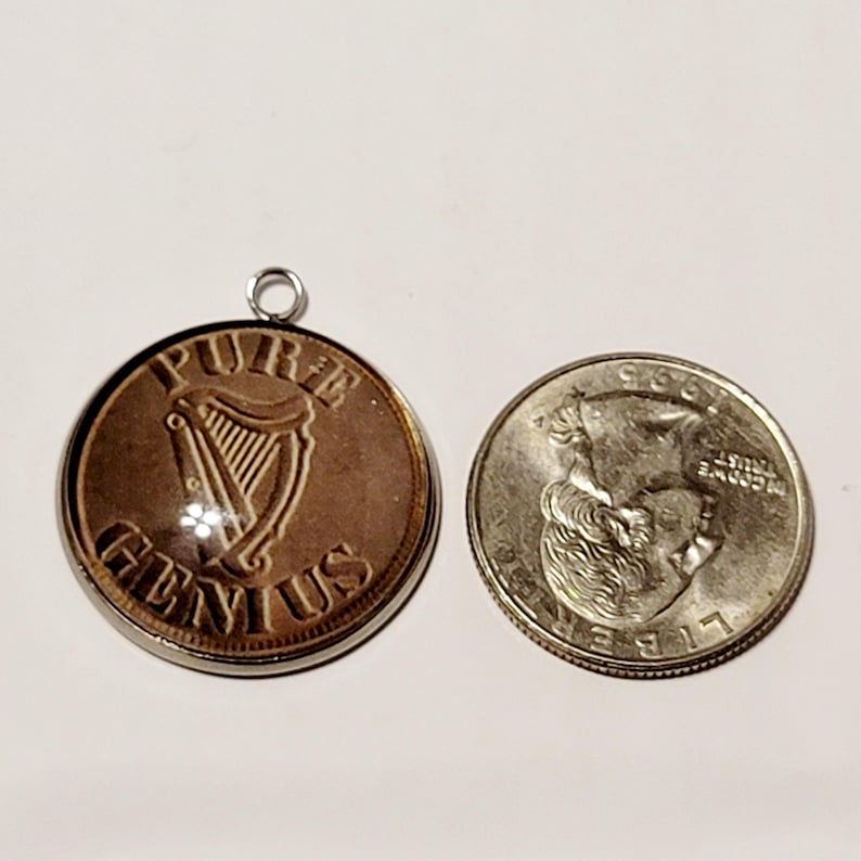 May include: A copper-colored pendant with the words "PURE GENIUS" and a harp design. Next to it is a silver quarter coin. The pendant has a silver loop at the top for hanging.