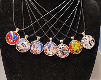 Puede incluir: Una colección de collares con colgantes redondos con varios diseños coloridos. Cada colgante está engastado en un bisel plateado y cuelga de un cordón negro. Los diseños incluyen texto e ilustraciones.