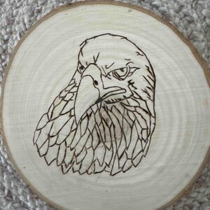 May include: A wood slice featuring a detailed pyrography design of an eagle's head. The eagle is depicted in profile, with intricate feather detailing and a focused expression. The natural wood grain provides a rustic backdrop.