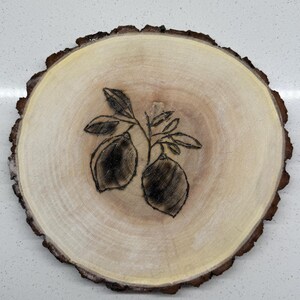 May include: A round wooden slice with a dark brown bark edge. The center features a pyrography design of two lemons and leaves. The wood has a light tan color with darker brown rings.