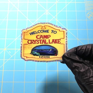May include: A yellow and blue sticker with a glittery border. The sticker says "Welcome to Camp Crystal Lake Est. 1935".