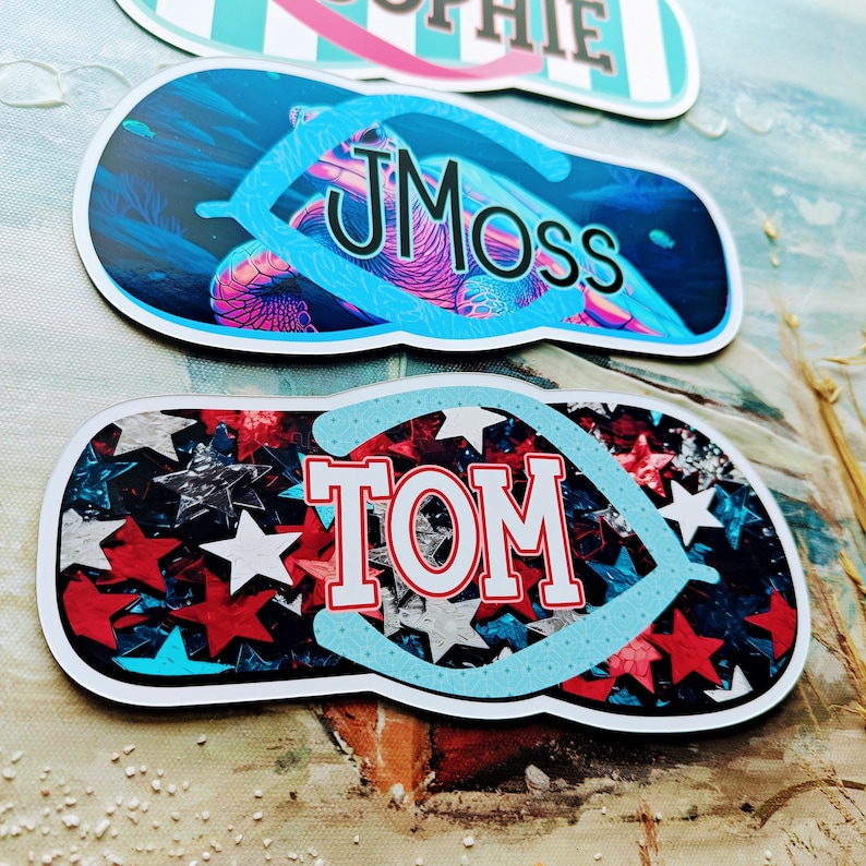 May include: Three flip-flop shaped stickers. One has the name "Sophie" in pink, another has "JMoss" over a sea turtle design, and the third has "TOM" over a red, white, and blue star pattern.