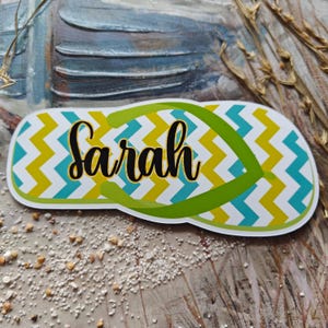 May include: A vibrant flip-flop shaped sticker. The design incorporates a white base with a zig-zag pattern in yellow and teal. The name "Sarah" is written in black script across the center, with a green strap.