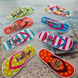 May include: A selection of personalised flip-flops with diverse patterns and names. Designs include stripes, zigzags, and floral prints in colours like green, red, blue, and yellow. Each flip-flop features a different name in a cursive font.