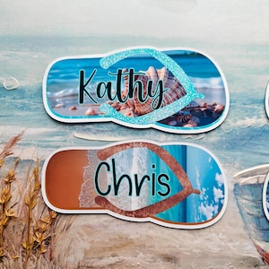 May include: Two personalised flip-flop-shaped name tags. One tag features the name "Kathy" in black script over a seashell image with a blue glitter border. The other tag has the name "Chris" over a beach scene.