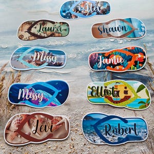 May include: A selection of flip-flop shaped stickers, each bearing a name such as "Alex" or "Jamie". The stickers showcase beach-themed designs, including seashells and ocean scenes. They are displayed on a sandy surface, evoking a seaside theme.
