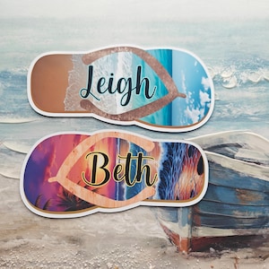 May include: Two personalised flip-flop shaped stickers. One features the name "Leigh" over a beach scene with blue water and sand. The other has the name "Beth" over a sunset beach scene with palm trees.