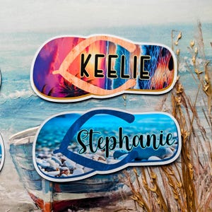 May include: Two personalised flip-flop-shaped stickers. One features the name "Keelie" with a sunset and palm tree design. The other has the name "Stephanie" with a beach pebble design. Both are on a painted ocean background.