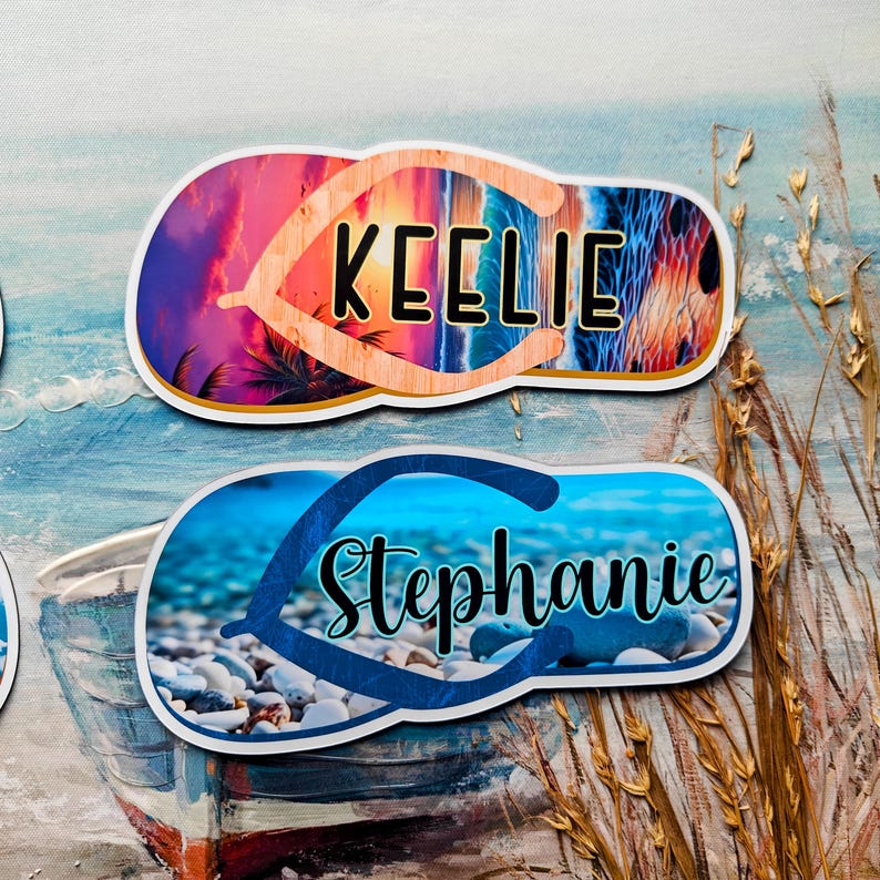 May include: Two personalised flip-flop-shaped stickers. One features the name "Keelie" with a sunset and palm tree design. The other has the name "Stephanie" with a beach pebble design. Both are on a painted ocean background.