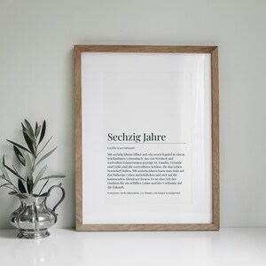 May include: A framed print with a white background and black text that reads "Sechzig Jahre" and a definition in German. The print is in a light wood frame and is sitting on a white surface. There is a small silver pitcher with green leaves in it to the left of the frame.