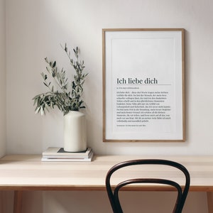 May include: A framed print with the German phrase "Ich liebe dich" meaning "I love you" in black text on a white background. The print is hanging on a white wall above a light wood desk with a vase of green branches and a stack of books.