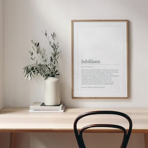 May include: A light wood desk with a framed print that reads "Jubil&auml;um" in black text. The print is on a white background. A vase with green branches is on the desk, along with a stack of books.